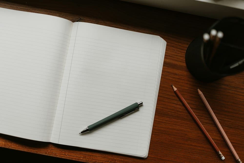 An open notebook with a pen and pencils on a desk.