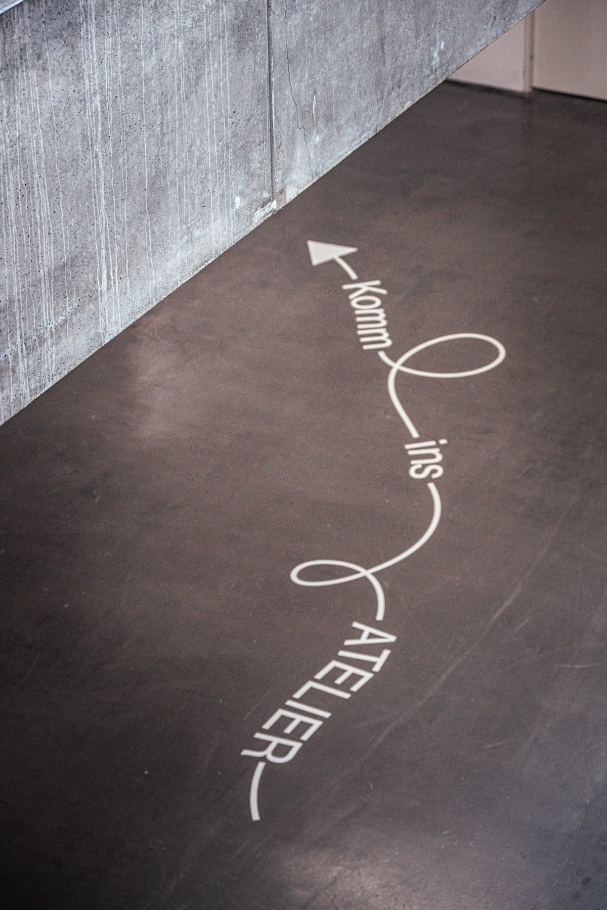 White looping lettering and an arrow on a dark gallery floor beside a concrete wall.